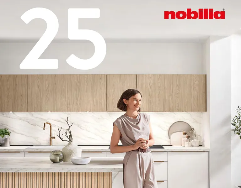 Helle Einbauküche mit glatten Holzoberschränken, weißer Arbeitsplatte in Marmoroptik und goldener Armatur, im Vordergrund eine Frau in beiger Kleidung mit Tasse in der Hand